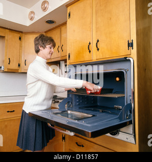 1960s 1970s SMILING HOUSEWIFE IN KITCHEN TAKING BAKING DISH OUT OF WALL MOUNTED OVEN Stock Photo