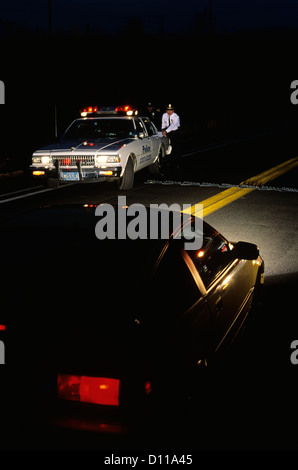 POLICE ROAD BLOCK TWO POLICE OFFICERS BEHIND PATROL CAR AND APPROACHING VEHICLE Stock Photo