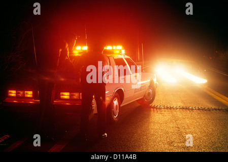 POLICE ROAD BLOCK TWO POLICE OFFICERS BEHIND PATROL CAR AND INCOMING CAR APPROACHING A SPIKE STRIP Stock Photo