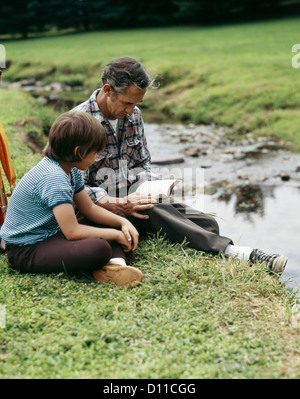 1970s MAN AND BOY FATHER AND SON OUTDOOR BY STREAM READING BIBLE Stock Photo