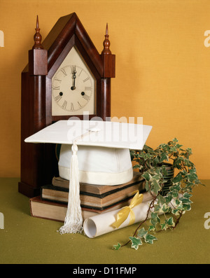 1970s WHITE GRADUATION MORTARBOARD CAP IVY PLANT DIPLOMA AND BOOKS WITH ANTIQUE CLOCK Stock Photo