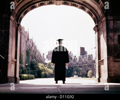 1960s 1960s BACK VIEW MALE GRADUATE WEARING CAP GOWN STANDING IN CAMPUS ARCHWAY Stock Photo