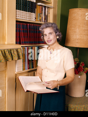 1960s MIDDLE AGED WOMAN READING HOLDING OPEN ENCYCLOPEDIA BOOK SMILING STANDING BY HOME LIBRARY LOOKING AT CAMERA Stock Photo