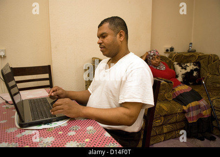 Husband who cares for his disabled wife at home (seated, amputated leg) uses laptop computer whilst wife watches TV Stock Photo