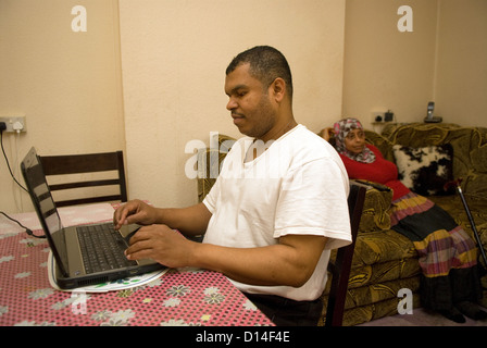 Husband who cares for his disabled wife at home (seated, amputated leg) uses laptop computer whilst wife watches TV Stock Photo