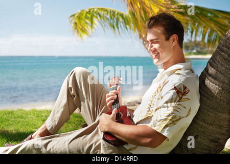 Hawaii, Oahu, Young Male With Vintage Aloha Shirt Relaxing On Palm Tree, Holding A Ukulele. Stock Photo
