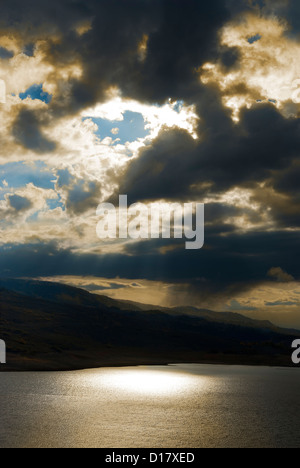 Dramatic stormy sky with sun and blue sky breaking through plus light filtering down to hills and a lake glistening below. Stock Photo