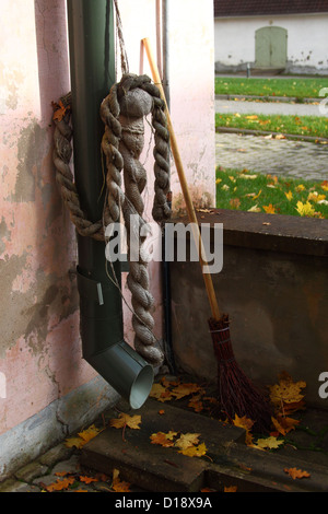 Autumn scene. Broom leaning against pink wall of the house near the drain pipe with rope hanging on it. Yellow fallen leaves. Stock Photo