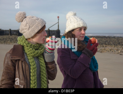 Two young women eating apples on beach in winter. Stock Photo