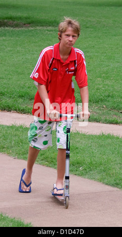Boy wearing bright red shirt and colourful shorts riding on scooter in parkland Stock Photo
