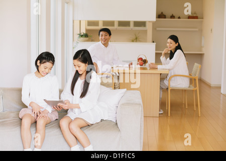 Sisters in pre-adolescent age using electronic tablet on the sofa with parents having coffee in the background Stock Photo