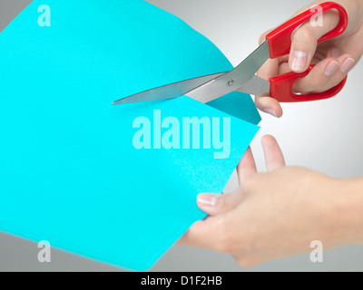 hands cutting a shape from a blue paper with a pair of red scissors on neutral grey background Stock Photo