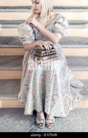 a woman in a floral dress is holding a pile of antique books on her lap Stock Photo