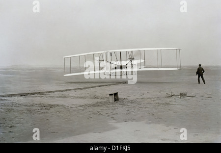 First successful flight of the Wright Flyer, by the Wright brothers. The machine traveled 120 ft in 12 seconds at 10:35 a.m. at Kitty Hawk, North Carolina. Orville Wright was at the controls of the machine, lying prone on the lower wing with his hips in the cradle which operated the wing-warping mechanism. Wilbur Wright ran alongside to balance the machine, and just released his hold on the forward upright of the right wing in the photo. The starting rail, the wing-rest, a coil box, and other items needed for flight preparation are visible behind the machine. This was considered the first sust Stock Photo