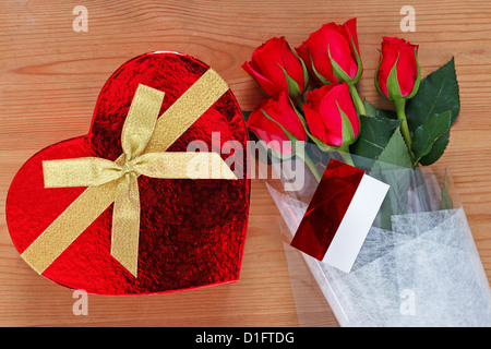 Chocolates in a heart shaped box and a bunch of red roses with card on an old wooden table. Stock Photo