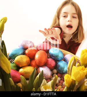 young girl reaching for coloured foil covered Easter eggs in basket surrounded by daffodils Stock Photo