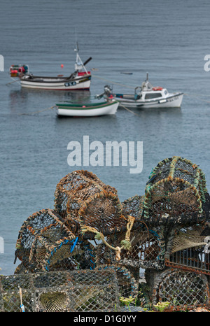 Lobster pots stacked on land, with three small fishing boats at anchor in harbour in background. Stock Photo