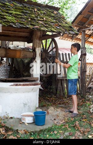 Schoolboy outdoor getting water from a vintage wooden well with pulley Stock Photo