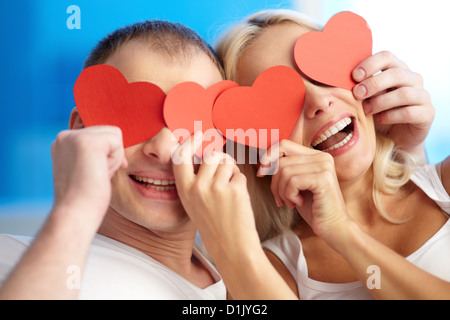 Portrait of joyful couple holding red paper hearts by their eyes and laughing Stock Photo