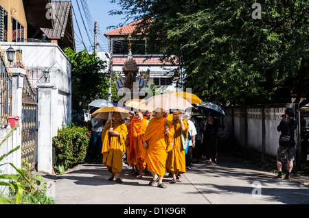 Buddhist monks wearing bright orange robes and carrying umbrellas for shade lead funeral procession with tall float on street Stock Photo