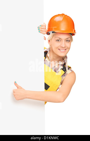 Female manual worker giving a thumb up and standing behind blank panel, isolated on white background Stock Photo