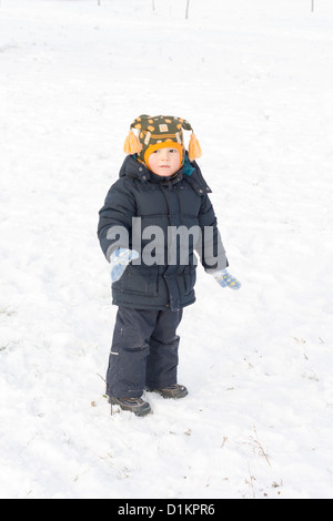 Adorable little boy standing in snow dressed in warm winter clothing with a hat with cute tassles looking at the camera Stock Photo