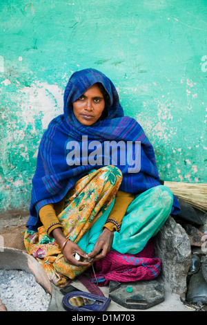 An Indian woman in a blue shawl sits outside her house against a green wall applying make up in the morning in Rajasthan India Stock Photo