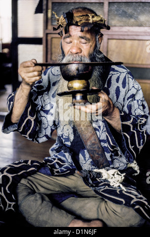 An elderly bearded Ainu man uses a wooden lifter to keep his mustache out of a drinking cup in Hokkaido, Japan, East Asia. Photographed in 1962. Stock Photo