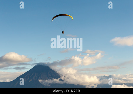A paraglider with a black and yellow canopy is suspended in mid-air against a clear blue sky, flying above a large, dark mountain peak partially shrouded in white clouds. The scene is set during Stock Photo