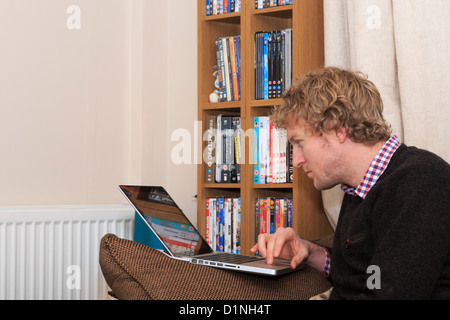 Young Millennial man squinting at a screen of a laptop computer resting on a chair arm in a living room at home. England UK Britain Stock Photo