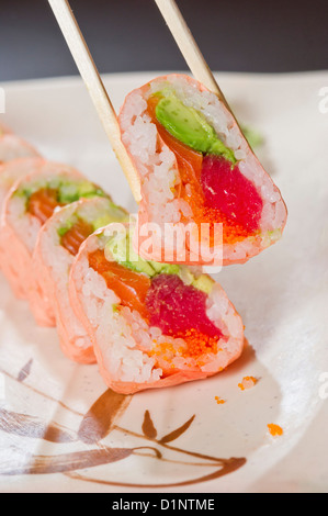 Chopsticks pick up a sushi roll with pink rice paper, tuna, salmon and avocado. Stock Photo