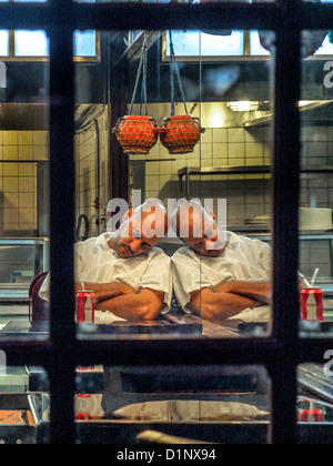 A tired African American cook is reflected in a wall mirror as he sleeps after restaurant closing late at night in Brooklyn. Stock Photo