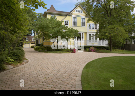 EXTERIORS Yellow clapboard Victorian with white trim curved cobblestone driveway three story home detached garage surrounded by Stock Photo