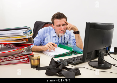 Confused accountant looking at some info on the display of his computer, surrounded by huge piles of documents. Stock Photo