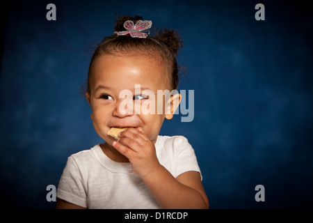 Cute studio portrait of a mixed race toddler girl eating a biscuit with a cheeky smile on her face Stock Photo