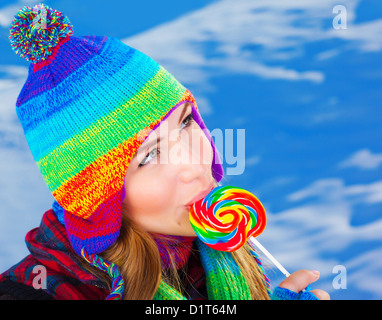 Image of attractive woman licking lollipop outdoors in wintertime, sweet colorful candy cane, snowy background, warm knitted hat Stock Photo