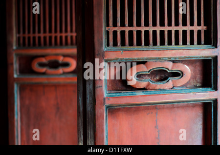 Temple Of Literature Carved Red Wooden Folding Doors Hanoi Vietnam | HANOI, Vietnam — Detail of a red lacquered folding door at the Temple of Literature, its carved rondel handle and slatted transom typical of traditional Vietnamese timber joinery. Founded in 1070 by Emperor Lý Thánh Tông, the temple complex added the adjoining Quốc Tử Giám (Imperial Academy) in 1076, making it Vietnam's first national university. The weathered lacquer and turquoise trim reflect generations of restoration across the Lê and Nguyễn dynasties. Stock Photo
