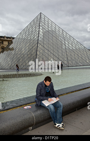 A young man sits looking at a map in front of the Louvre glass pyramid Paris, Ile de la Cite, France Stock Photo