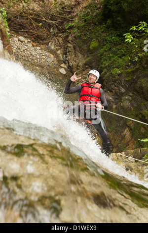 A man is rappelling down a rocky waterfall while wearing a white helmet, red life vest, and black wetsuit. He is holding a rope with one hand and making a hand gesture with the other, smiling. Water Stock Photo