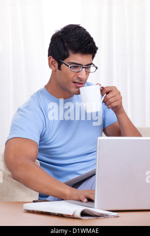 Young man taking a sip of coffee while operating laptop Stock Photo