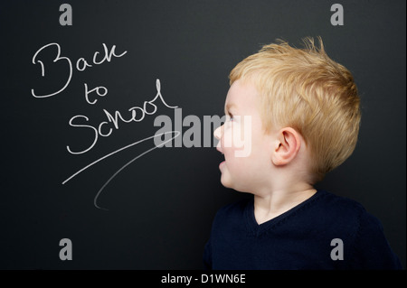 Smart young boy wearing a navy blue jumper stood in front of a blackboard with Back to School written in chalk