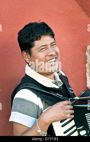 blind accordionist smiles as he stands outside on sidewalk against beautiful red wall talking with friend Oaxaca Mexico Stock Photo
