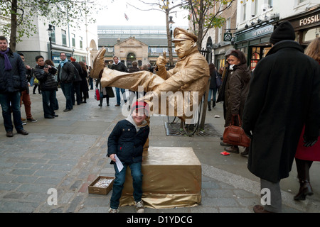 A young boy in crowd street posing watching a man street performer painted with gold paint balancing in Covent Garden, London England UK   KATHY DEWITT Stock Photo