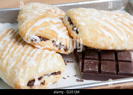 Three golden and flaky chocolate filled, iced croissants on a baking pan with wax paper, and squares and crumbles of chocolate. Stock Photo