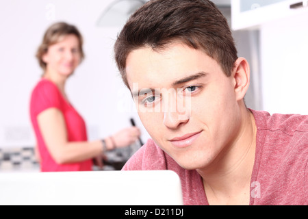 Young male using a laptop, a woman is in the background cooking. Stock Photo