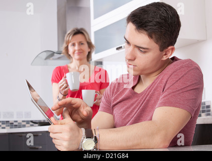 Young man using a digital tablet looking at the screen or monitor. Situated in a modern kitchen a woman holding two mugs smiles. Stock Photo