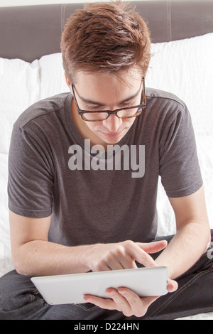 Young male wearing glasses looking at the screen of his digital tablet, sitting on his bed. Stock Photo
