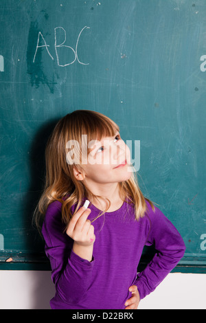 Young girl standing in front of chalkboard with letters of the alphabet and holding stick of chalk in hand Stock Photo