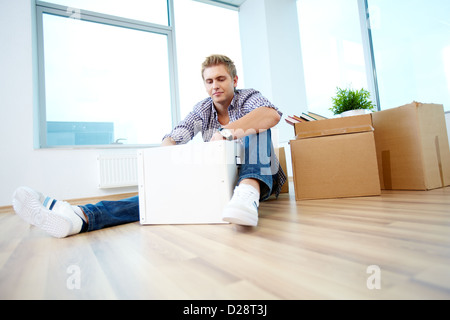 A young guy sitting on the floor of new house and unpacking box Stock Photo
