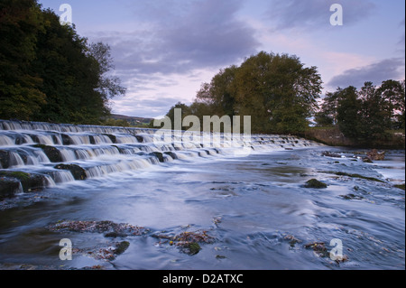 Scenic, evening, rural landscape of water flowing & tumbling over & down weir steps - River Wharfe, Burley in Wharfedale, Yorkshire, England, UK. Stock Photo